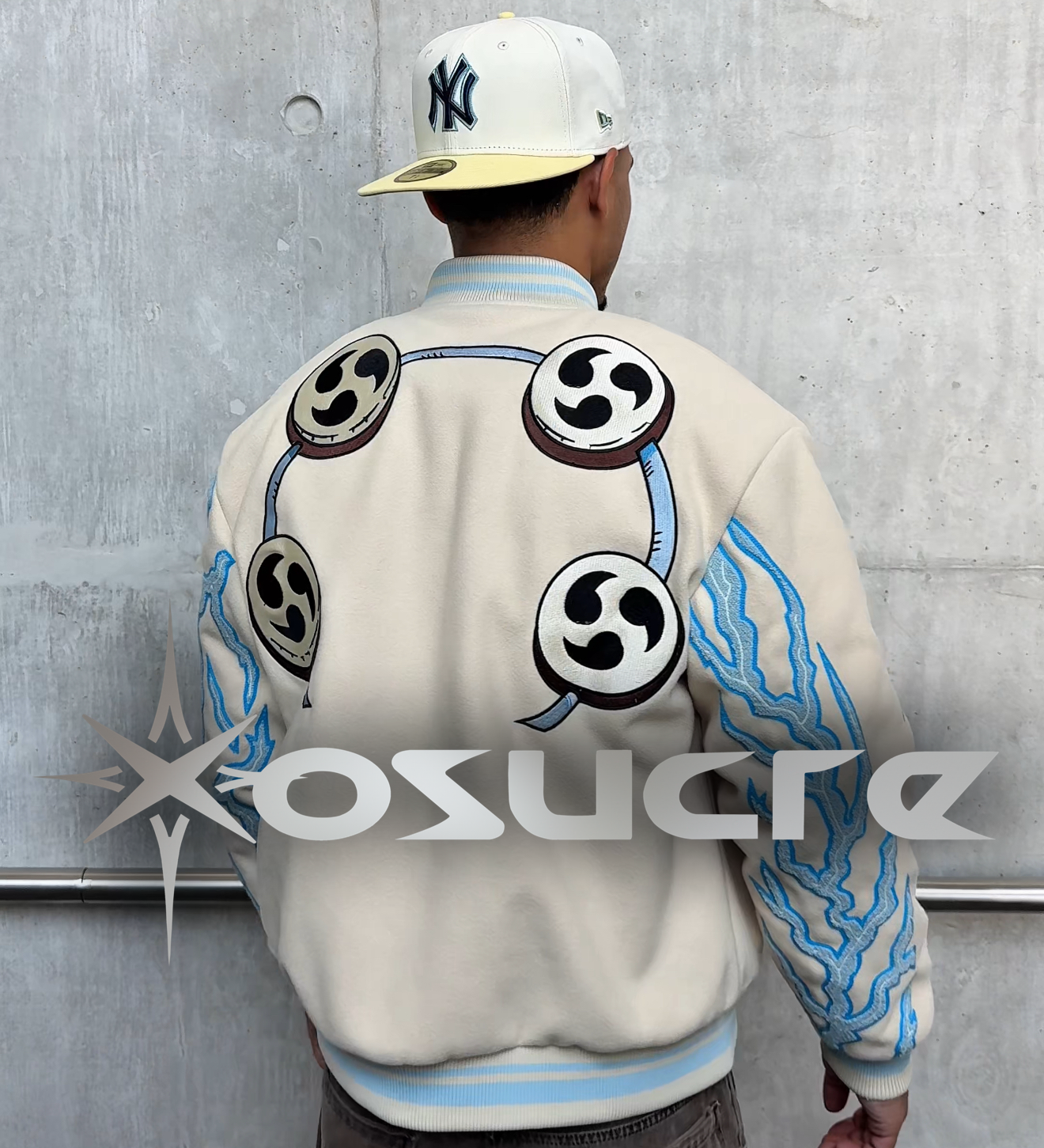 Person wearing a cream-colored jacket with black and blue designs, standing against a gray wall.